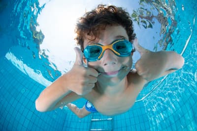 Child smiling and giving two thumbs up while swimming underwater.